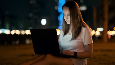 A-woman-software-developer-sits-in-a-Park-on-a-summer-night-in-the-city-and-writes-code-looking-at-a-laptop-screen.-Print-the-article-on-a-laptop.-Blogger-replies-to-posts
